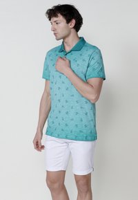 Türkiz színű pólóing, texturált mintával és kontrasztos gallérral, fehér rövidnadrággal párosítva. Rövid ujjú és laza szabású.