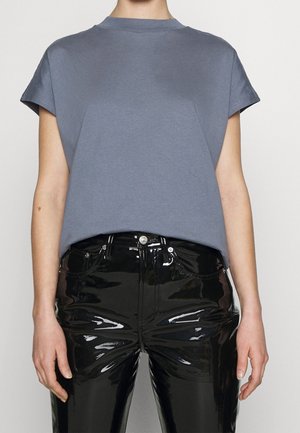 Woman wearing a loose gray sleeveless top paired with shiny black patent leather pants, standing against a plain light background.