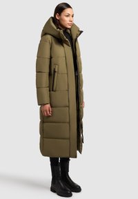 Lång, olivgrön puffjacka med hög krage, quiltat mönster, sidofickor och dragkedja framtill. Bärs över svarta byxor och stövlar.