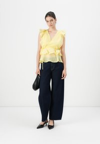 Blusa amarilla transparente con mangas acampanadas, diseño en capas, combinada con jeans de mezclilla oscuros de pierna ancha y tacones negros de punta.