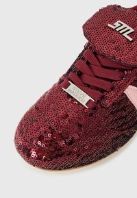 Red sequined sneaker with textured surface, featuring black laces and a pink accent. Includes a metallic brand tag on the side.