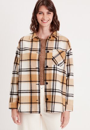 Geruite shirt in geel, wit en zwart met een knoopsluiting aan de voorkant, een borstzak en een relaxed fit. Gedragen over een bruine trui.