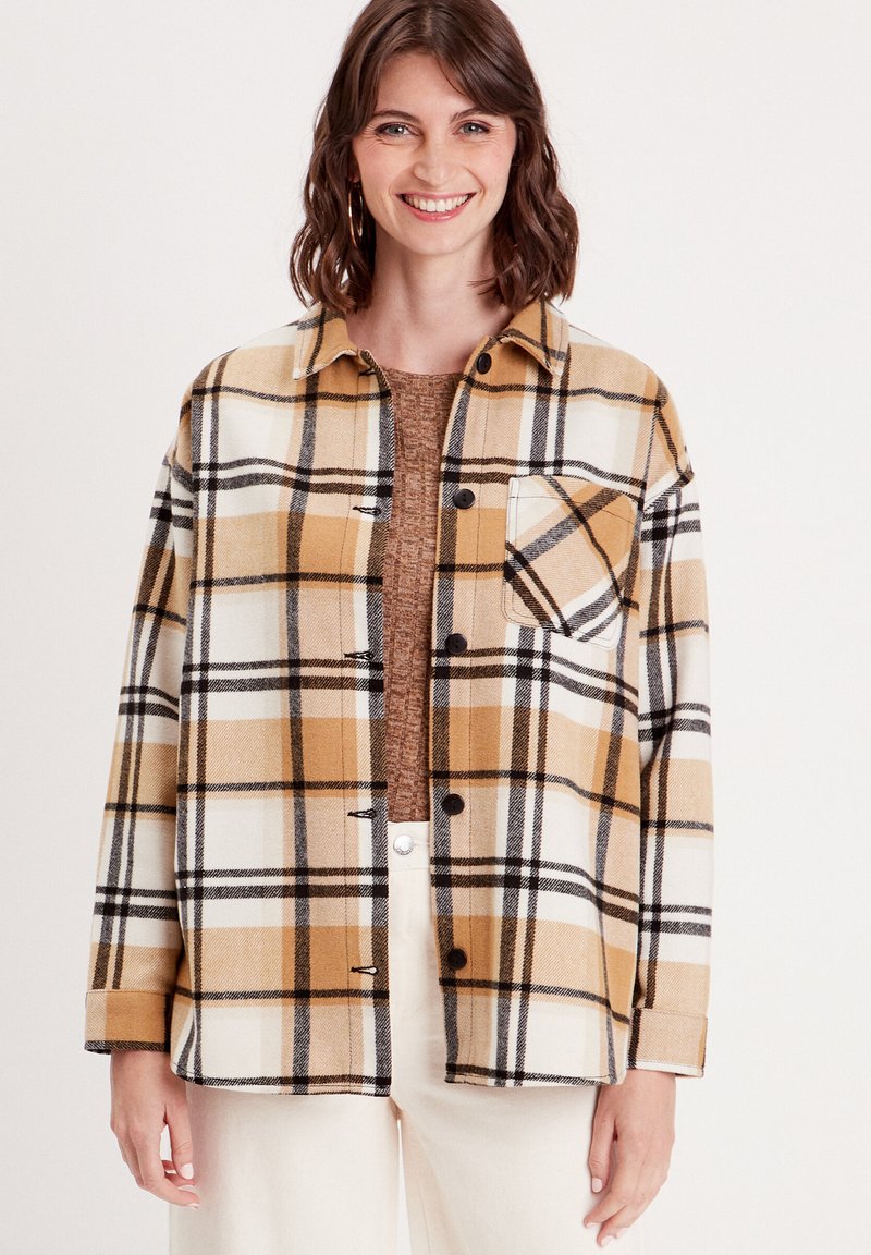Camisa de cuadros en amarillo, blanco y negro con cierre de botones, un bolsillo en el pecho y un corte relajado. Usada sobre un suéter marrón.