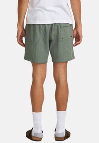 Shorts rayés verts avec une poche arrière, fabriqués en tissu léger. Présentent des rayures jaunes et une coupe décontractée. Portés avec des chaussettes blanches et des sandales.