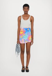 Top alb, fustă mini florală coloreată cu modele roz, albastru și galben și geantă cu franjuri gri. Pantofi plate negri cu design cu barete.