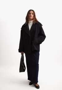 Cappotto di lana nera doppiopetto con ampi risvolti, abbinato a una lunga gonna in denim scuro e scarpe nere flat, mentre tiene una borsa nera.