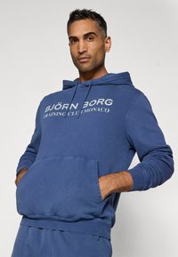 Kék kapucnis pulóver pamut keverékből, elülső zsebbel, állítható zsinóros kapucnival és "BJÖRN BORG TRAINING CLUB MONACO" logónyomattal.