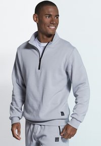 Light grey zip-up sweatshirt made of soft fabric, featuring a high collar, long sleeves, and a small logo patch at the hem.