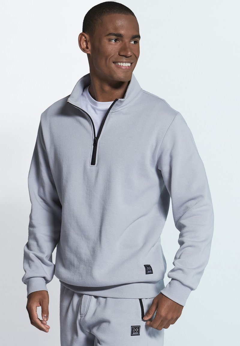 Light grey zip-up sweatshirt made of soft fabric, featuring a high collar, long sleeves, and a small logo patch at the hem.