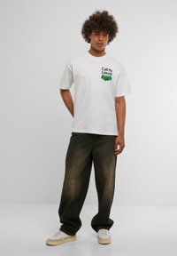 Hvid bomulds t-shirt med teksten "Ring til min advokat" og grafik på brystet, parret med mørke, løstsiddende jeans og sneaks.