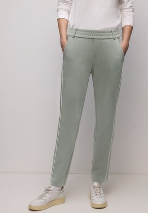 Osoba oblečená v světle zelených kalhotách s bílými pruhy po stranách, bílém dlouhém rukávu a bílých teniskách, stojící s rukama v kapsách.