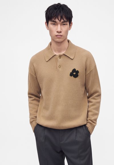 Homme portant un pull en maille beige avec col et détail fleur noire sur la poitrine, associé à un pantalon gris foncé, debout les mains dans les poches.