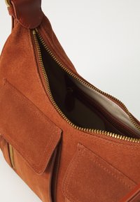 Wildlederhandtasche in Rostfarbe mit goldfarbenem Reißverschluss. Verfügt über zwei Fronttaschen, Lederdetails und ein geräumiges Innenfach.