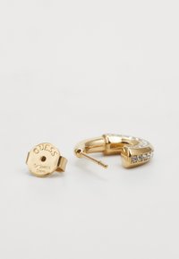Guld hoopörhänge med en böjd design. Ena sidan pryds av klara rhinstenar, och baksidan visar det ingraverade varumärket och materialet.