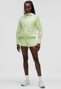 Helle grüne leichte Jacke mit Kapuze und elastischen Manschetten, kombiniert mit passenden Shorts. Das Model trägt weiße Sneakers und Knöchelsocken.