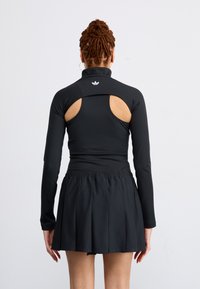 Woman with braided hair wearing a black long-sleeve top with shoulder cutouts and a pleated black skirt, standing against a plain white background.