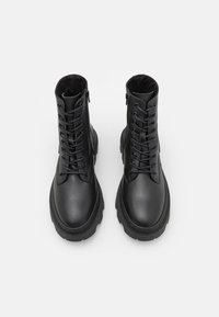 Une paire de bottes de combat noires en cuir à lacets, avec des fermetures éclair latérales, des semelles épaisses et une finition texturée sur fond blanc.