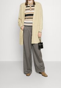 Hellbeige Strickjacke, gestreiftes Oberteil in Beige, Schwarz und Weiß, graue weite Hose und Schlangenmustermuster Stiefeletten. Kleine schwarze Handtasche.