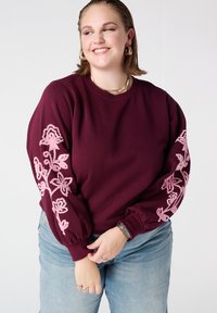 Bordeauxrode trui met roze bloemenborduursel op de mouwen, ronde halslijn en elastische boorden. Gecombineerd met lichtblauwe spijkerbroek.