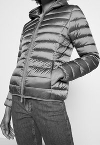 Zilveren pufferjack met een ritssluiting aan de voorkant, horizontaal doorgestikt ontwerp, capuchon en elastische manchetten, gecombineerd met donkere jeans. Soepele textuur.