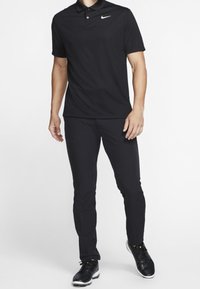 Muž v černém polokošili Nike, černých kalhotách a černých teniskách stojí na bílém pozadí.