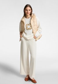Gilet trapuntato beige sopra una maglia bianca con un motivo circolare dorato, abbinato a pantaloni beige a gamba larga e scarpe marroni slip-on.