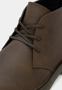 Rudi odiniai kulkšnies batai su apvalia nosimi, tekstūruota paviršiaus apdaila, raišteliais ir subtiliais siūlių akcentais palei kraštus.