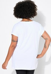 T-shirt blanc à manches courtes, avec une coupe décontractée, une texture lisse et un col rond. Tatouages visibles sur les bras.