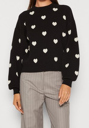 Maglione nero con motivi a cuore crema, scollatura rotonda e maniche oversize. Abbinato a pantaloni grigi a righe. Materiale morbido e testurizzato.