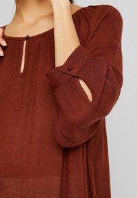 Femme ajustant la manche d'un chemisier à manches longues couleur rouille avec poignet boutonné et petit détail en forme de trou de serrure au niveau de l'encolure.