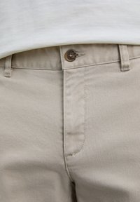 Pantalones de algodón beige con un botón marrón abrochado en la cintura, combinados con una camisa blanca metida por encima de la cinturilla.