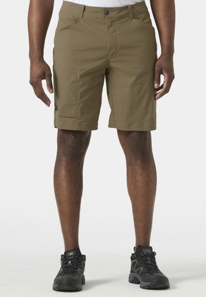 Hombre vistiendo pantalones cortos color verde oliva hasta la rodilla y zapatos de senderismo negros, posando contra un fondo claro y liso.