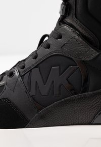 Černá teniska s texturovanou kůží, hladkou tkaninou, tónovaným logem značky a černými tkaničkami. Design zahrnuje výraznou střední podrážku a ozdobné prvky.