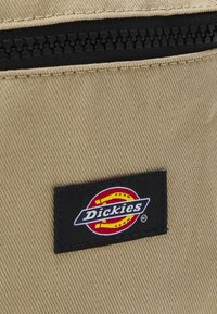 Khaki szövet táska fekete cipzárral a tetején. Jellemzője egy téglalap alakú fekete címke fehér szöveggel és egy piros és sárga kör alakú logóval.