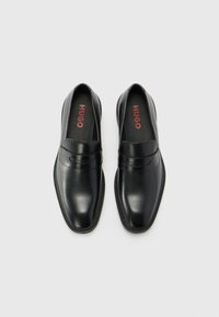 Mustad musta nahast loafers sileda viimistlusega, millel on struktureeritud varvas, peened õmblusdetailid ning bränditud tallad punase tekstiga.