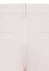 Pantalon rose pâle avec fermeture éclair, doté de deux poches arrière et d'une texture lisse et mate. Coutures visibles le long des coutures.