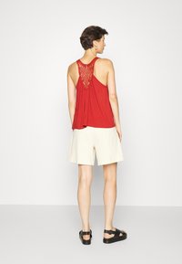 Top vermelho sem mangas com um detalhe nas costas em crochê, corte relaxado e textura suave, combinado com calções cor creme e sandálias pretas.