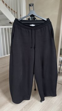 Zwarte sweatpants met wijde pijpen en een taillekoord, hangend aan een zwarte hanger binnen, nabij een houten vloer en een wit hek.