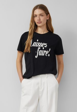 Kobieta z długimi blond włosami, ubrana w czarną koszulkę z napisem "Laissez-faire!" i białe spodnie z wysokim stanem, stojąca na szarym tle.