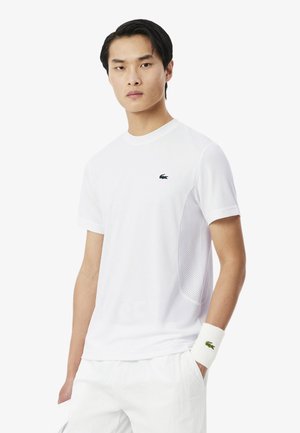 Giovane uomo che indossa una maglietta bianca Lacoste con pannelli laterali in mesh, pantaloncini bianchi e un polsino bianco con il logo del coccodrillo verde.