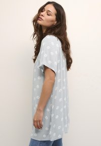 Blouse bleu clair avec un motif de fleurs blanches, manches courtes et une coupe fluide. Tissu doux avec une surface texturée et une coupe décontractée.
