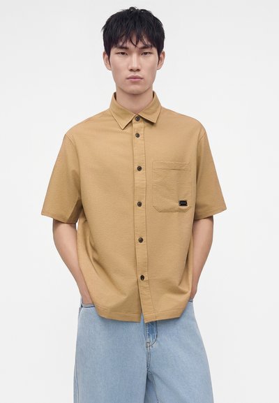 Jeune homme aux cheveux noirs portant une chemise beige à manches courtes et un jean bleu clair, debout les mains dans les poches devant un fond uni.