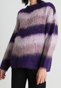 Pull rayé violet avec différentes nuances de violet et de rose. Fabriqué à partir d'un matériau duveteux et texturé. Présente un large col rond et de longues manches.