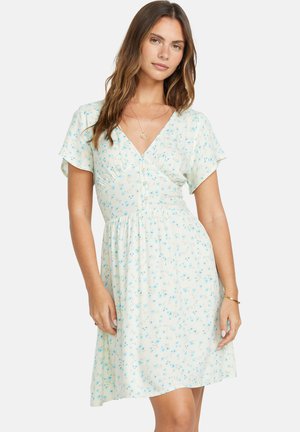 Vrouw draagt een lichtgekleurde jurk met korte mouwen en een blauw bloemmotief, een V-hals, knopen aan de voorkant, en een gouden ketting en armband.