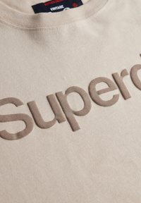 Beige bomulds sweatshirt med en hævet brun "Superdry" logo på forsiden. Klassisk crew neckline med ribbede detaljer.