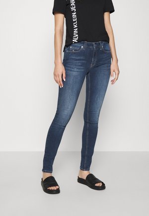 Mujer con jeans ajustados azul oscuro, camiseta negra de Calvin Klein y sandalias negras de tira, de pie frente a un fondo claro.