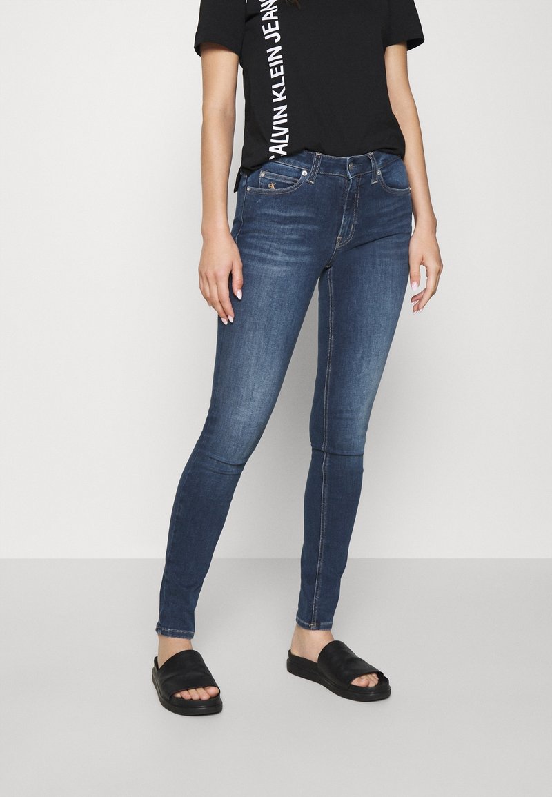 Mujer con jeans ajustados azul oscuro, camiseta negra de Calvin Klein y sandalias negras de tira, de pie frente a un fondo claro.