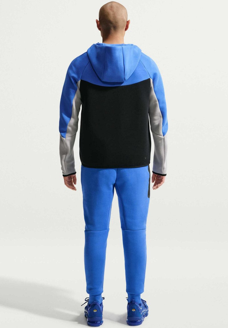 Homme portant un sweat à capuche bicolore avec des pans bleus, noirs et gris, un jogging bleu et des baskets bleues assorties, vu de dos.