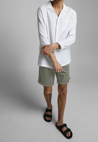 Persona vestita con una camicia bianca a maniche lunghe, pantaloni corti verde oliva e sandali neri con fibbia, in piedi su uno sfondo grigio semplice.