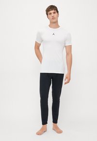 Homme debout pieds nus, portant une chemise blanche à manches courtes avec un logo et un pantalon noir ajusté, sur un fond blanc uni.